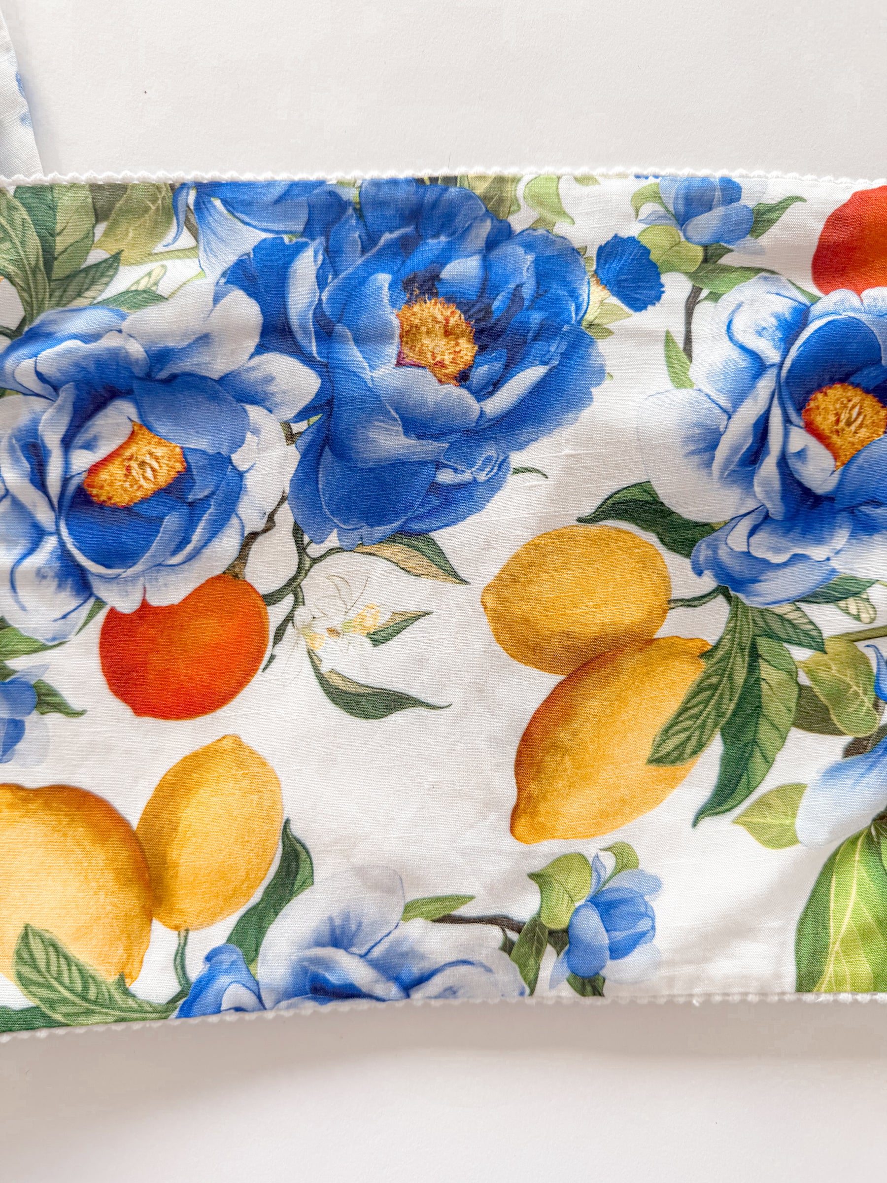 A close-up of a fabric product, likely a pouch or bag, featuring a watercolor-style floral print with blue peonies, yellow lemons, and green leaves on a white background.