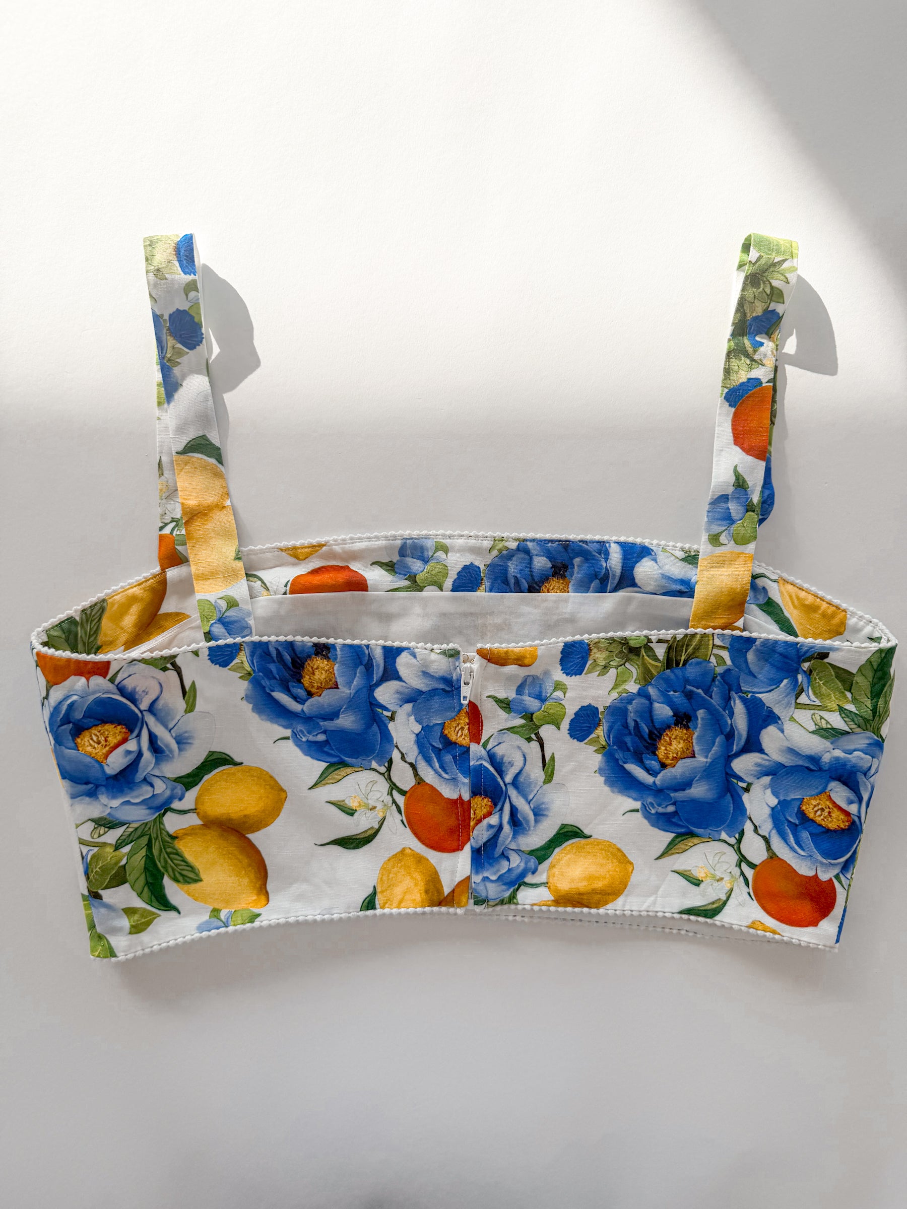 A white bikini top with a vibrant floral and citrus print featuring blue flowers, yellow lemons, and orange oranges, displayed against a plain white background.