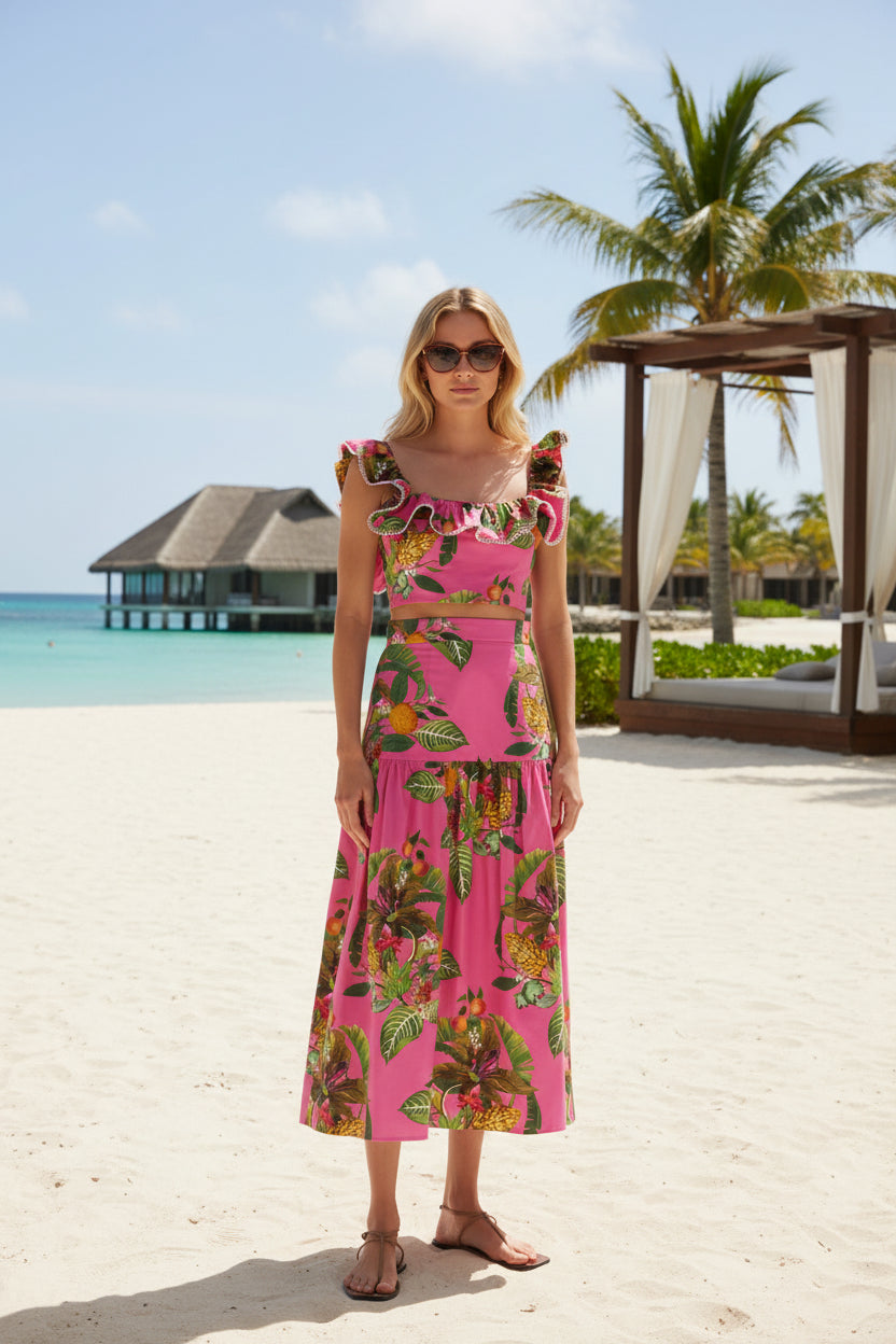 A model wearing a bright pink two-piece set featuring a ruffled crop top and a matching tiered midi skirt, both printed with a tropical green and yellow leaf pattern, set against a plain white studio background.