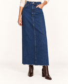 A dark blue denim maxi skirt with a raw, frayed hem, modeled with a cream long-Agolde Hilla Denim Maxi Skirt sleeve top and dark brown heeled ankle boots against a plain white studio background.