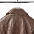 A close-up of a brown leather jacket hanging on a white hanger against a white background, showing the collar and a small square brand patch on the back.