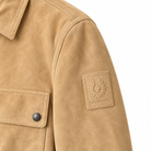 Belstaff Men's Suede Tour Overshirt Sand Close-up of a tan suede jacket sleeve featuring a square tan Belsenstaff patch on the upper arm and a matching pocket with a dark metal snap on the chest.