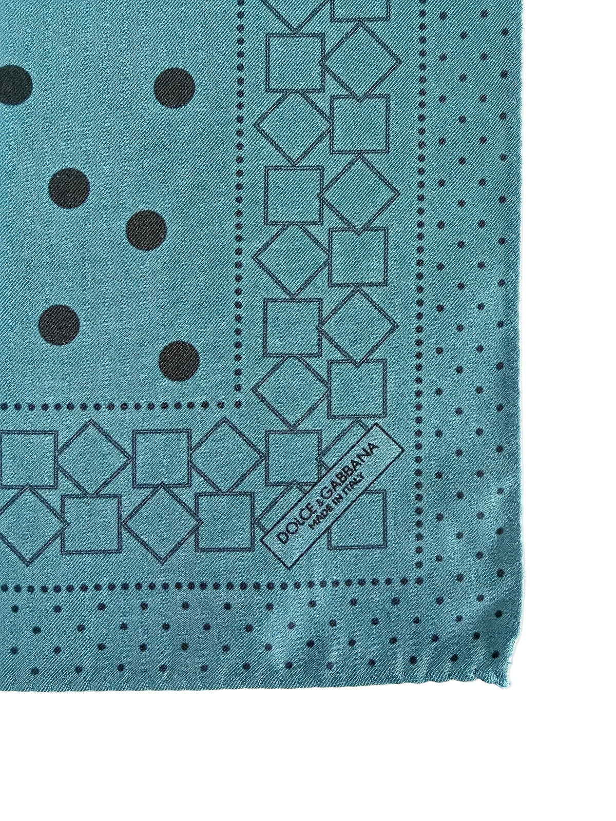 A close-up of a Dolce & Gabbana scarf in a dusty teal blue color, featuring black geometric patterns, polka dots, and a white brand label stitched onto one corner.