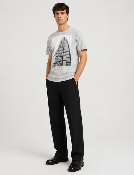 A man modeling a light gray crewneck t-shirt with a black and white architectural Dries Van Noten Grey Hertz Tower T-Shirt building graphic on the front, paired with black trousers and black leather shoes, against a plain light gray studio background.