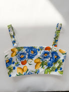 A white strap top featuring a vibrant floral and fruit print with blue flowers, yellow lemons, and orange citrus fruits, displayed flat against a white background.