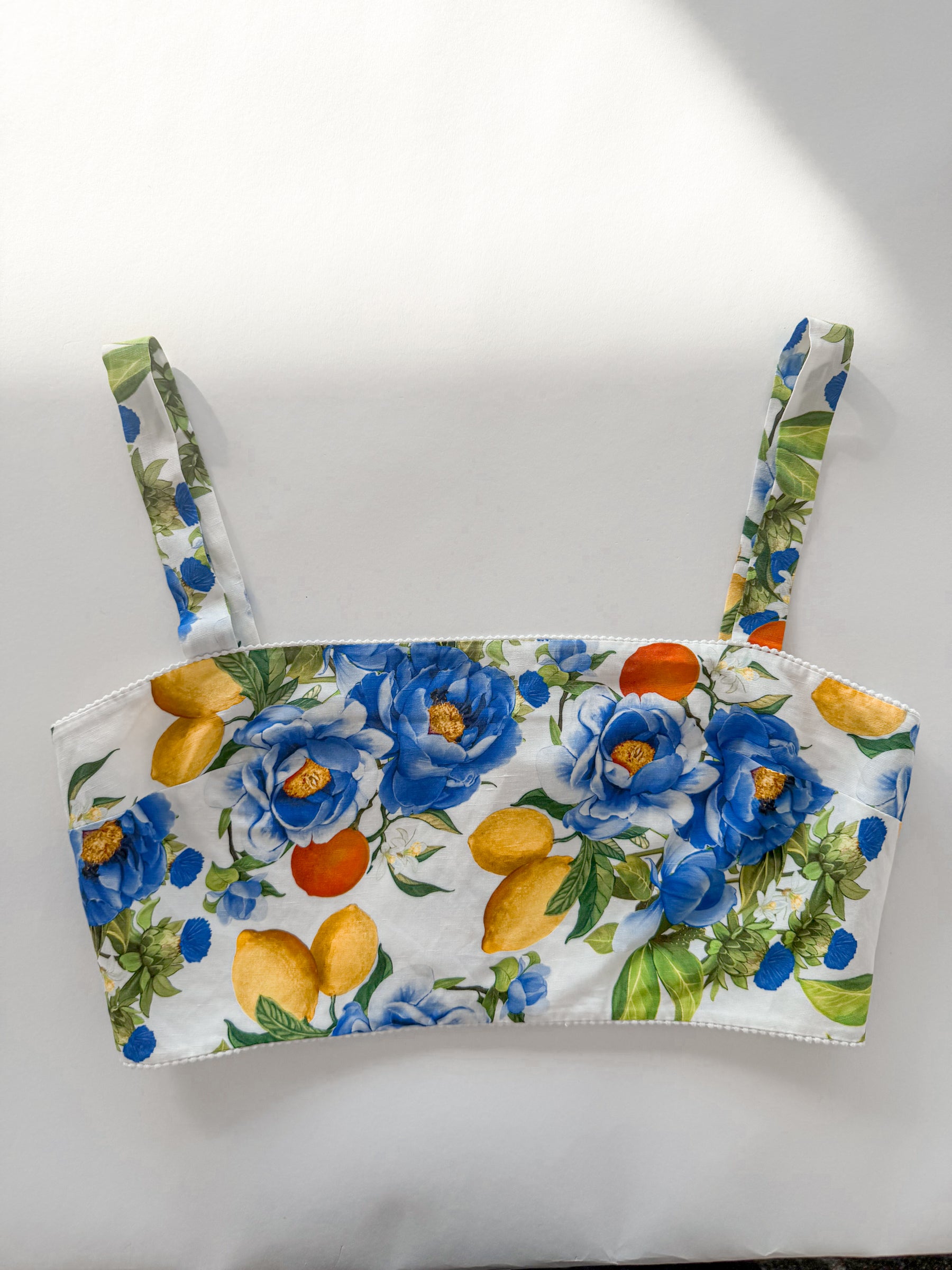 A white strap top featuring a vibrant floral and fruit print with blue flowers, yellow lemons, and orange citrus fruits, displayed flat against a white background.