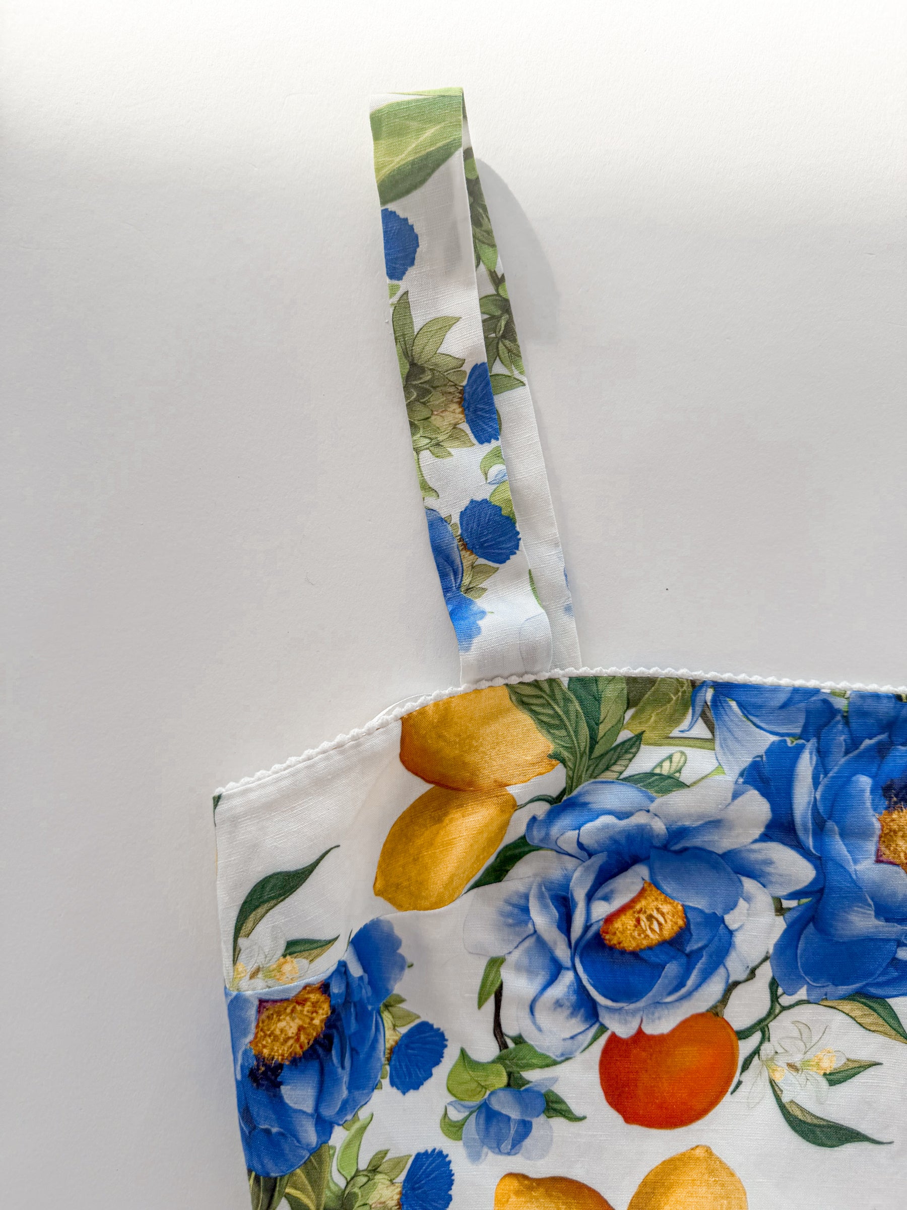 A close-up of a fabric tote bag featuring a colorful floral print with blue flowers, yellow-orange fruits, and green leaves on a white background.