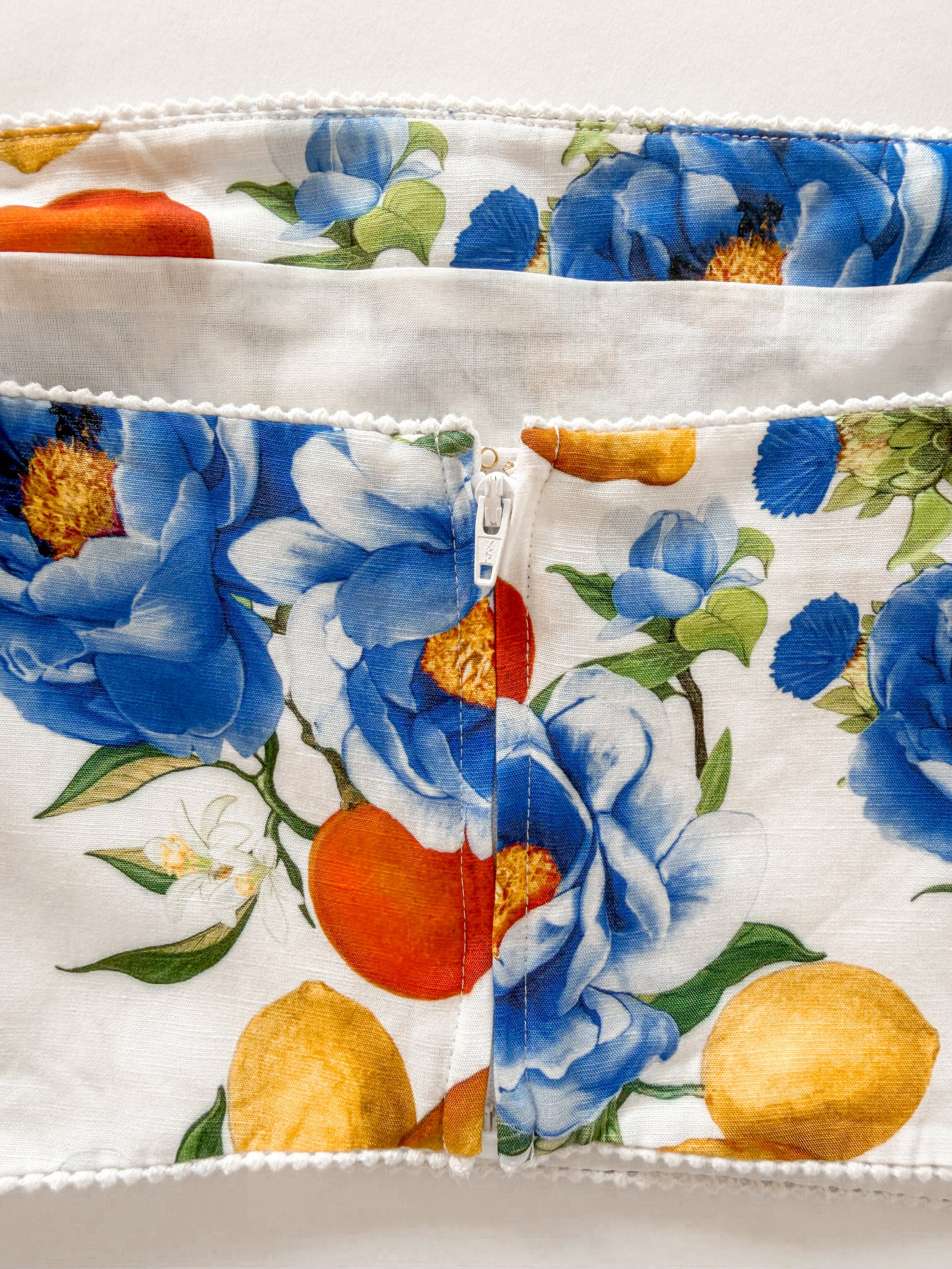 Close-up of a fabric item with a white zipper closure and a colorful floral print featuring large blue flowers, orange and yellow fruits, and green leaves on a white background.