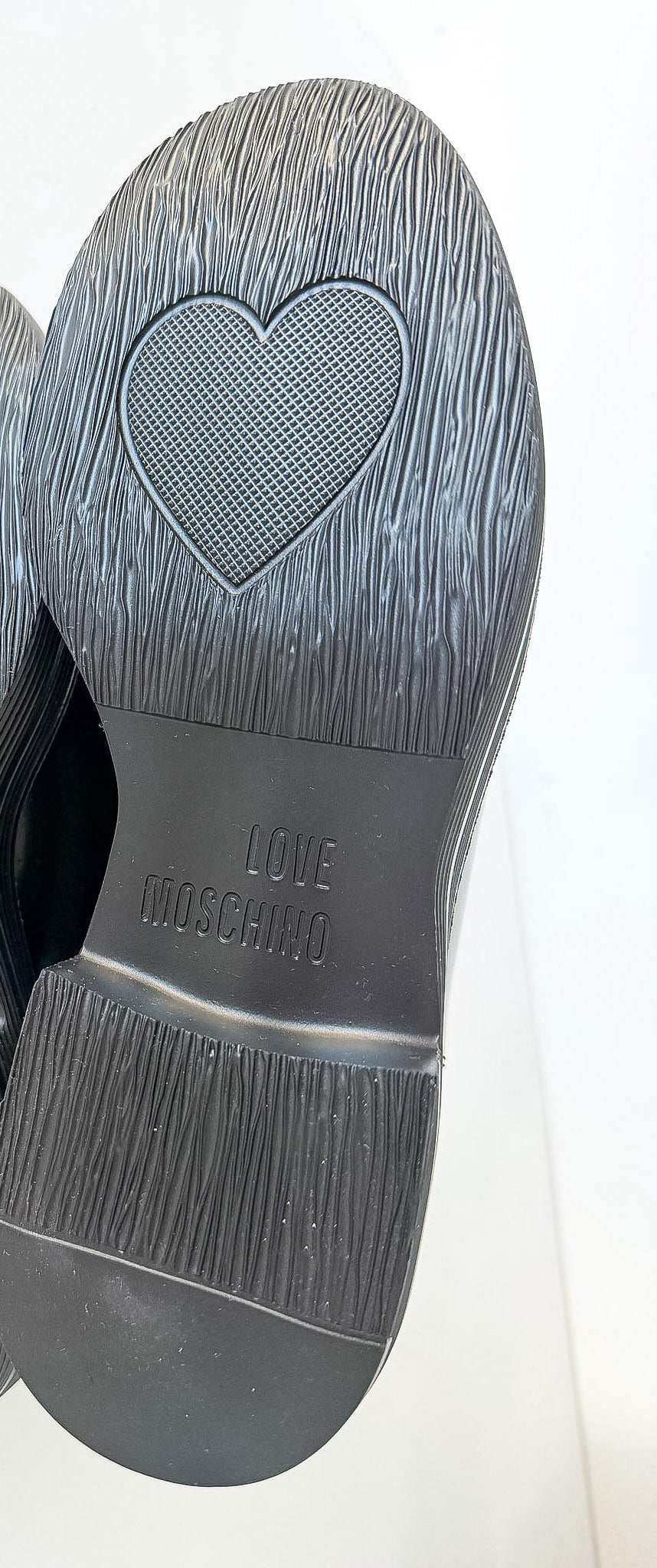 The soles of a pair of black shoes, showing a textured rubber tread with a heart-shaped grip pattern and the brand name 'LOVE MOSCHINO' embossed on the heel.