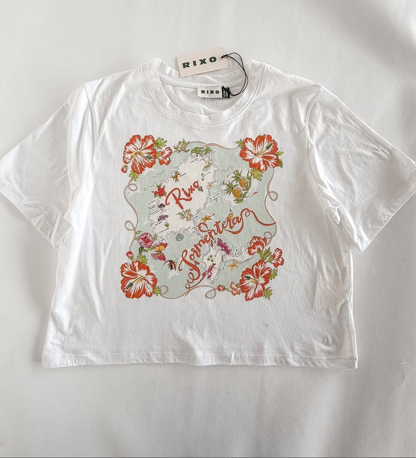 A white short-sleeve crop top t-shirt by RIXO, featuring a colorful graphic print on the front with red hibiscus flowers, green leaves, and a central illustration. The brand tag 'RIXO' is visible at the collar.