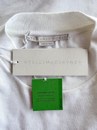 A close-up of a white crewneck sweatshirt laid flat on a white fabric background, showing the interior neck opening, a white brand hangtag, a green material tag, and a small sewn-in size/brand label. The embossed brand name STELLAMCCARTNEY is visible on the hangtag.