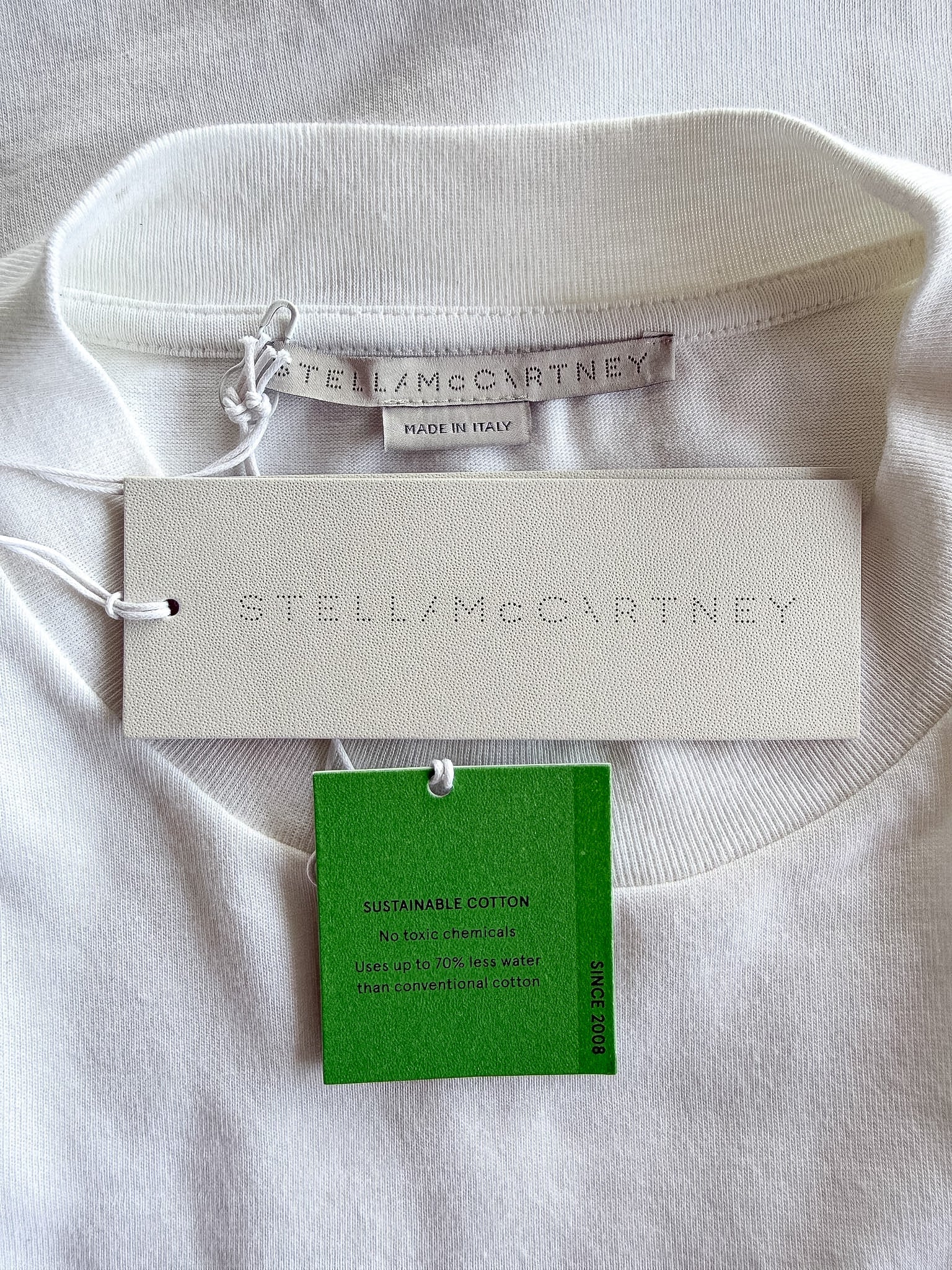 A close-up of a white crewneck sweatshirt laid flat on a white fabric background, showing the interior neck opening, a white brand hangtag, a green material tag, and a small sewn-in size/brand label. The embossed brand name STELLAMCCARTNEY is visible on the hangtag.