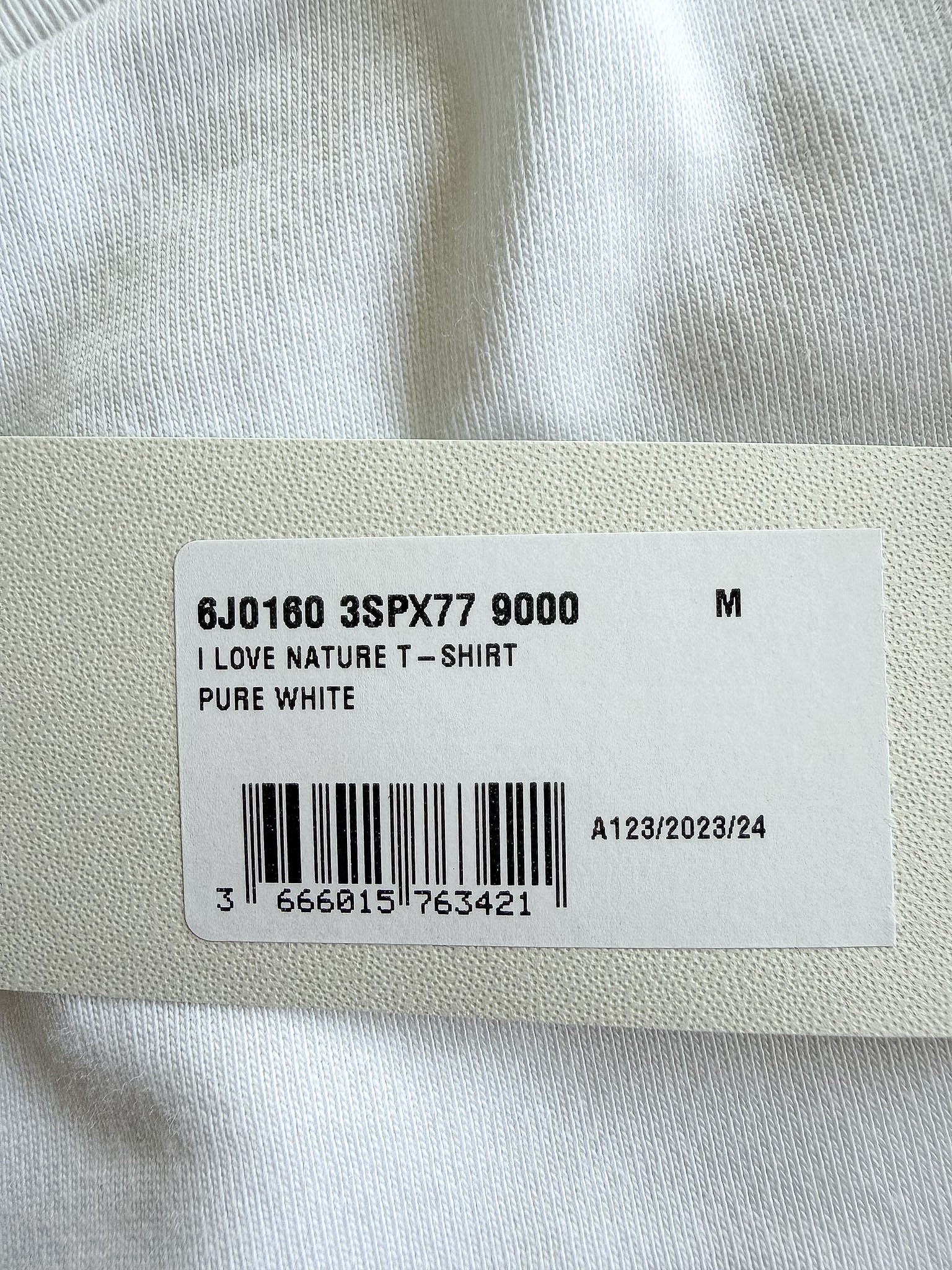 A close-up of a white garment tag attached to a white textured fabric item, showing a barcode and printed text including 'PURE WHITE' and size information.