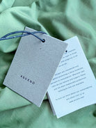 A gray textured paper tag with the brand name 'ASCENO' printed in black, attached to a blue cord, laid on a light green fabric background next to a printed card.