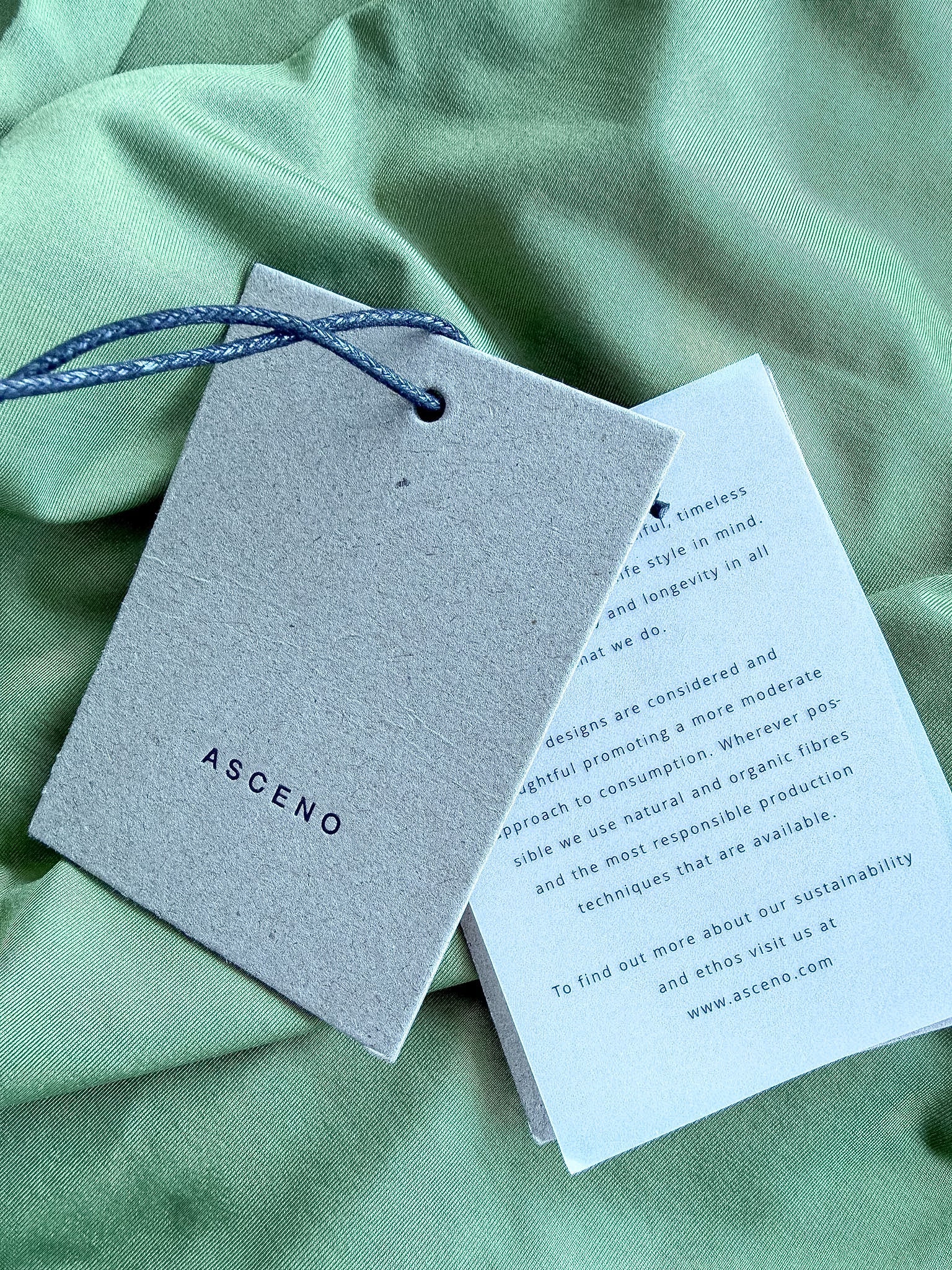 A gray textured paper tag with the brand name 'ASCENO' printed in black, attached to a blue cord, laid on a light green fabric background next to a printed card.