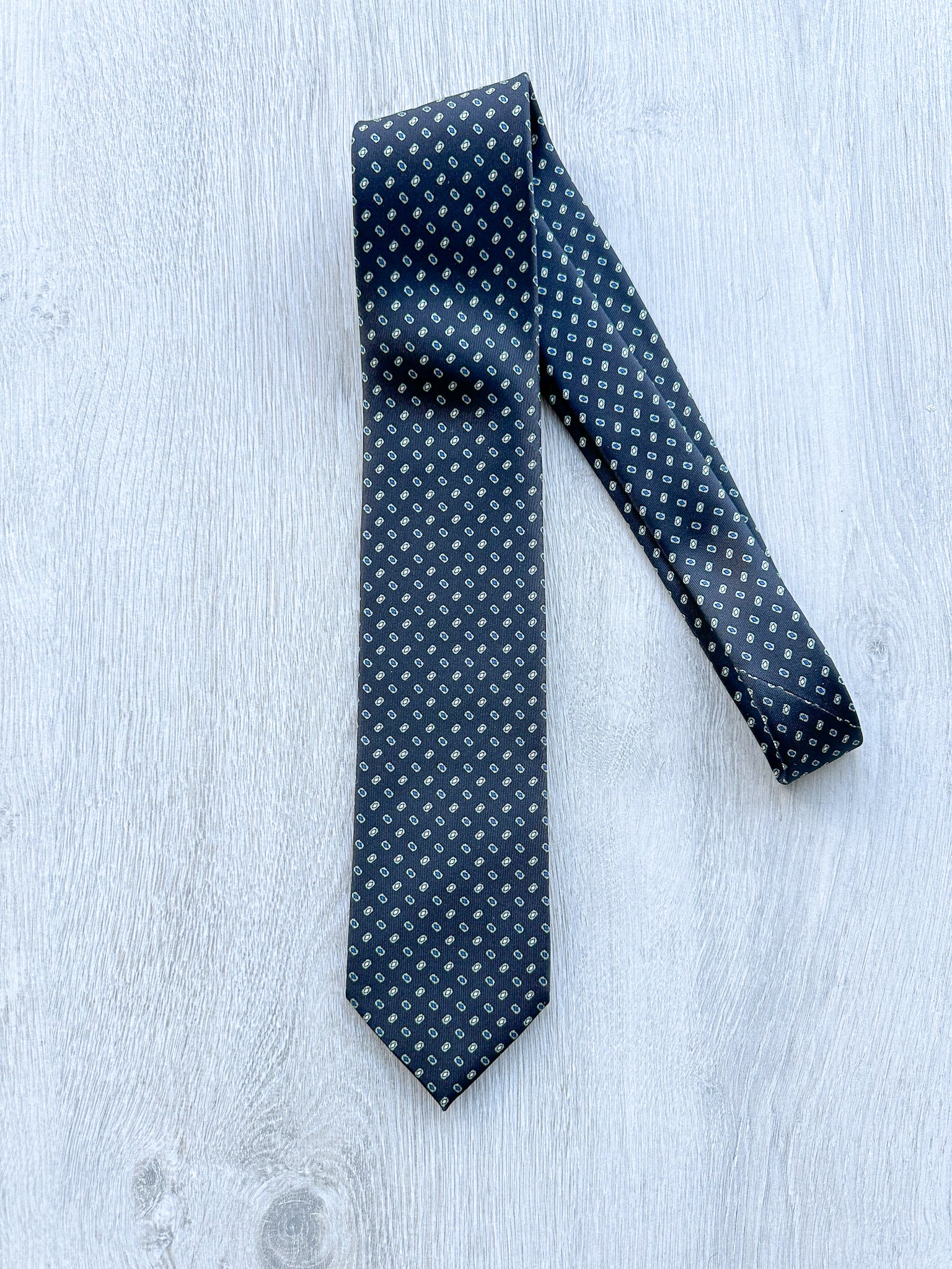 A black silk tie with teal square diamond print, laid out on a wooden surface.