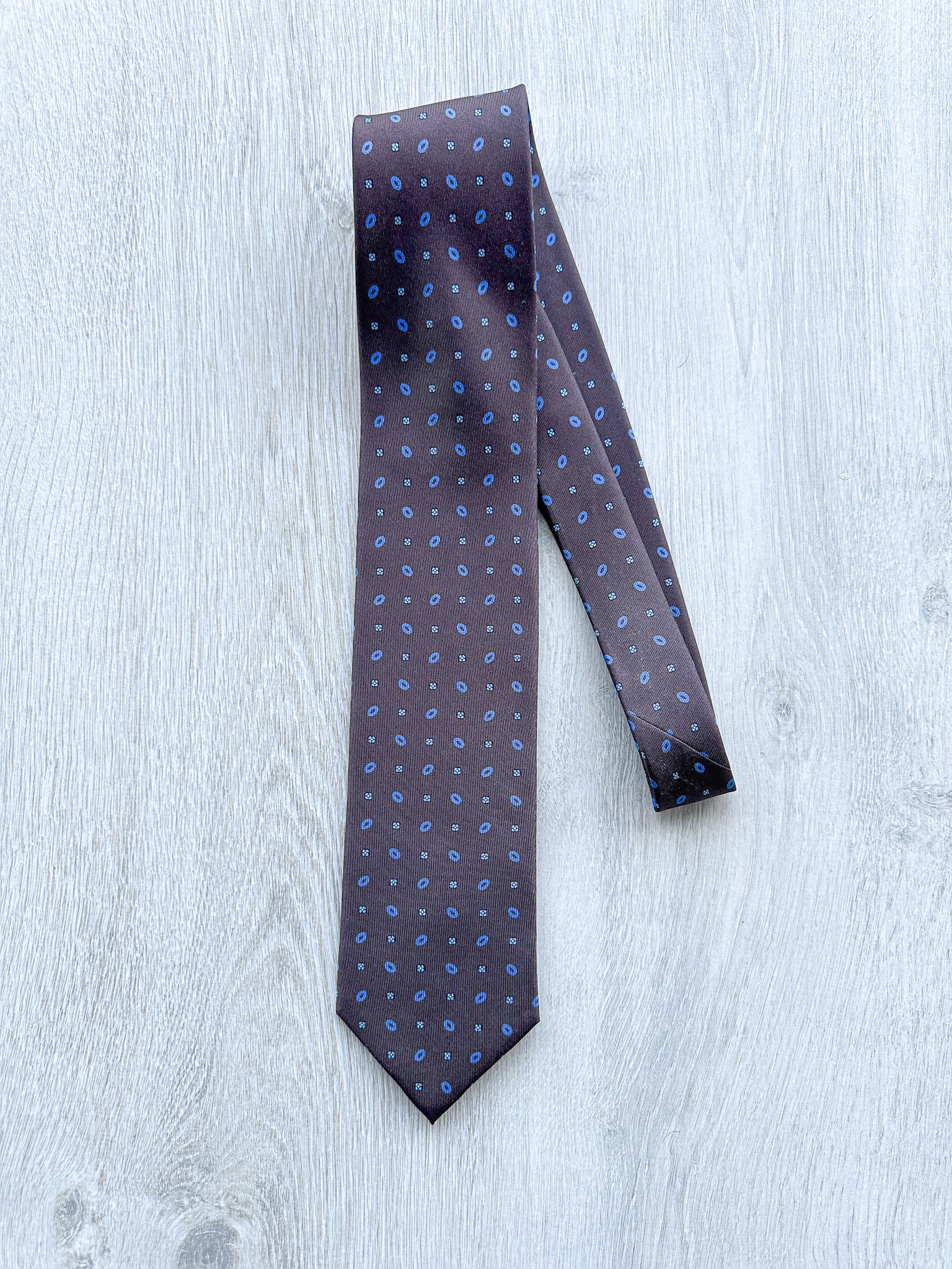 A chocolate brown silk tie with a blue circle and diamond floral print, displayed on a wooden surface.