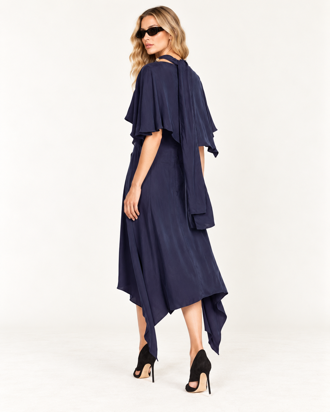 J.W. Anderson Navy Cape Style Draped Dress (minor flaw) A woman modeling a navy blue midi dress with layered flutter sleeves and an asymmetrical draped hem, styled with black sunglasses and black high-heeled sandals against a plain white studio background.