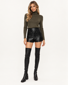 Maje Black Tweed Vinyl High Waist Shorts A woman modeling a dark olive green long-sleeve turtleneck sweater paired with black high-waisted faux-croc shorts and black over-the-knee boots, shot against a plain white studio background.