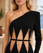 A close-up of a model wearing a black one-shoulder long-sleeve dress with geometric cutouts and a waist tie, set against a softly blurred outdoor background.