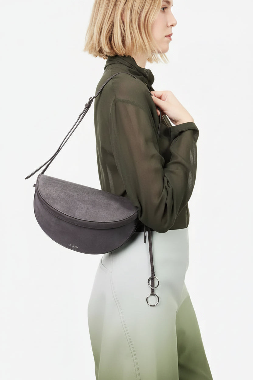A model shown from the side wearing a Alaia dark gray suede crescent-shaped shoulder bag with a thin black strap, styled with an olive green sheer long-sleeve top and a light green skirt against a plain white background.
