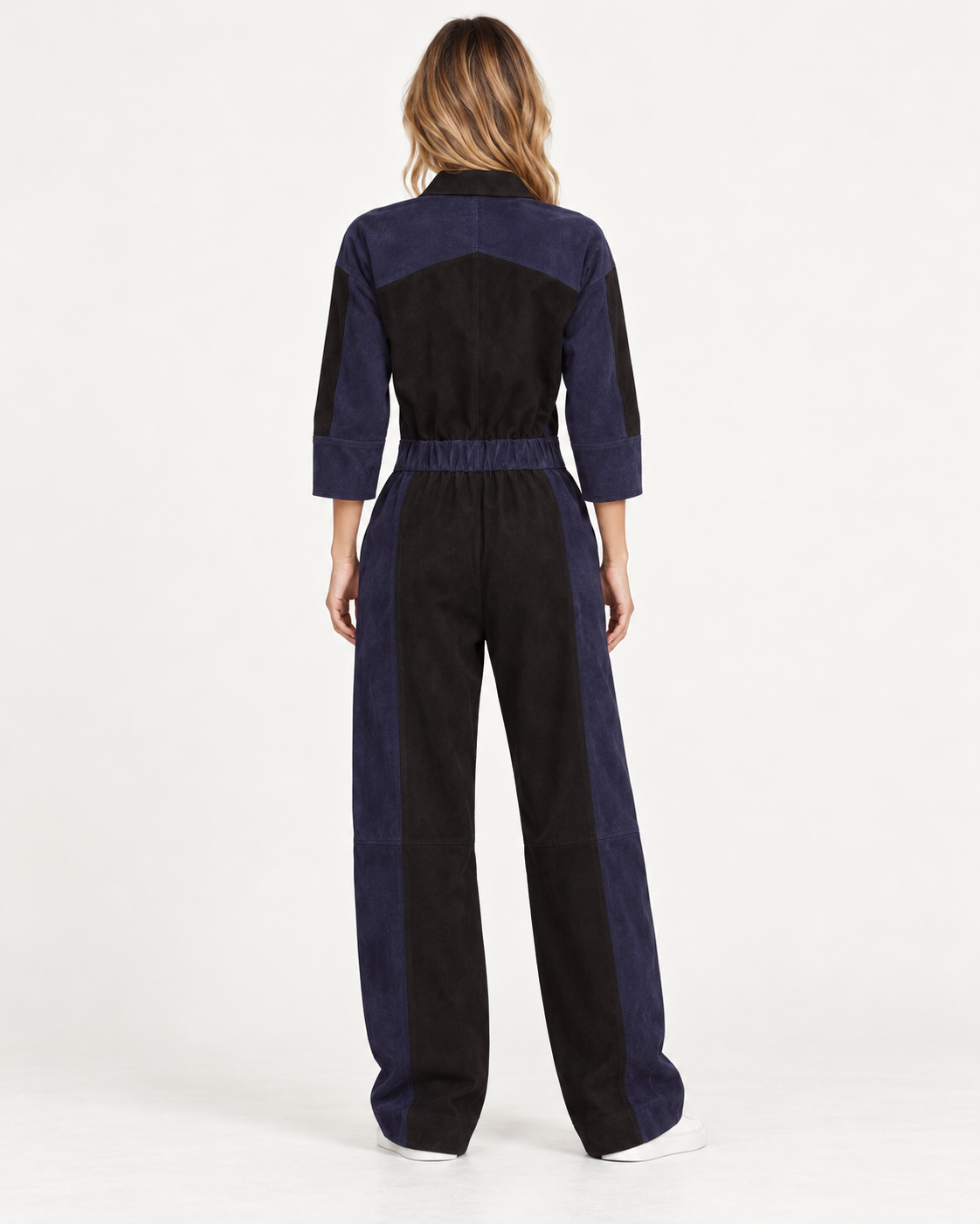 Raey Nappa Suede Leather Patchwork Suede Jumpsuit A model viewed from the back wearing a color-blocked jumpsuit made of navy blue and black fabrics, set against a plain white studio background.