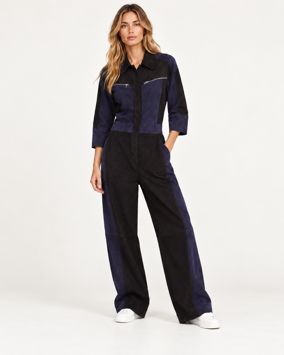 Raey Nappa Suede Leather Patchwork Suede Jumpsuit A woman modeling a dark navy and black color-blocked jumpsuit with wide-leg pants, a collared shirt-style top, and silver zipper accents on the chest, paired with white sneakers against a plain white studio background.