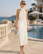 A woman stands by a resort pool wearing a sleeveless, high-neck, midi-length white sequined dress with vertical textured stripes. She is accessorized with a small metallic handbag and sunglasses.