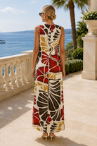 A woman stands on a sunlit terrace overlooking the sea, photographed from the back. She is wearing a sleeveless maxi dress with a bold abstract floral pattern in red, black, mustard yellow, and cream.