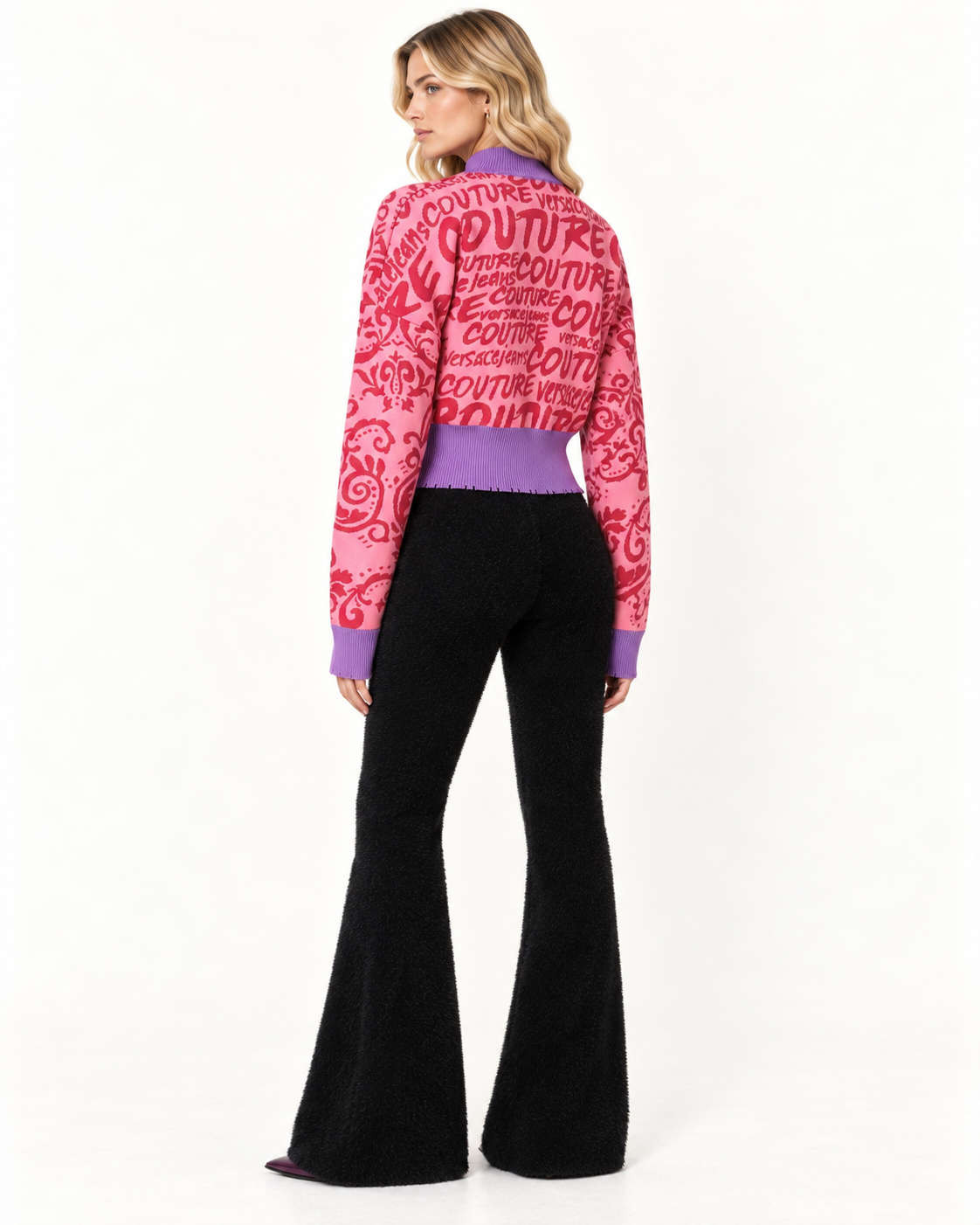 Versace Jeans Couture Pink Knitted Allover Logo Jumper A model shown from the back wearing a pink and magenta cropped knit cardigan with a 'COUTURE' pattern, paired with black flared trousers, against a plain white studio background.