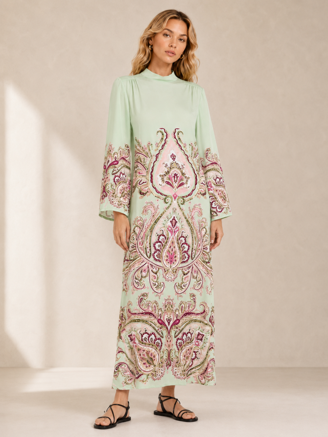 A woman modeling a hayely mezies long-sleeve maxi dress with a mint green base and a large pink, purple, and beige paisley pattern across the front and sleeves, set against a neutral beige studio background.