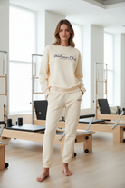 Person wearing a cream-colored 'Wellness Club' sporty & rich sweatshirt with a reformer pilates studio background