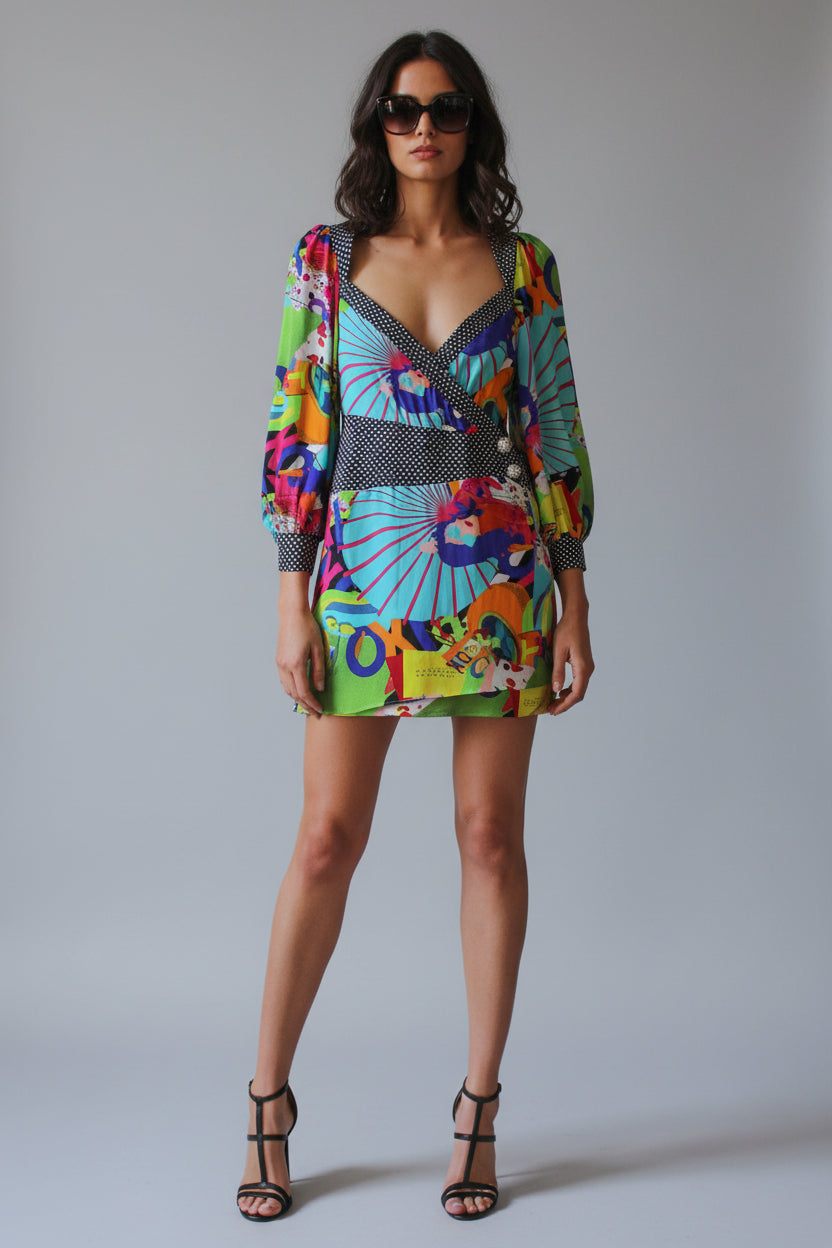 A woman stands indoors wearing a short, long-sleeve wrap dress with a bold multicolor abstract print and a navy-and-white striped waist detail. 