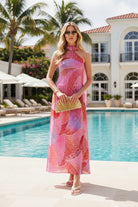 A model wears a vibrant pink and red patterned halter-neck maxi dress with a matching neck scarf, holding a gold folding fan. The dress features an abstract, flowing wave pattern in shades of pink, red, and orange.