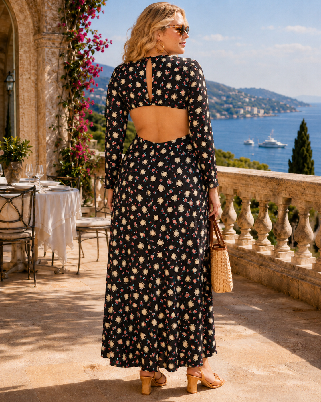 A woman stands on a sunlit terrace overlooking a blue sea and hills, wearing a long-sleeve black maxi dress with a cream and beige floral print and an open back. She is seen from behind, holding a woven straw bag.