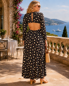A woman stands on a sunlit terrace overlooking a blue sea and hills, wearing a long-sleeve black maxi dress with a cream and beige floral print and an open back. She is seen from behind, holding a woven straw bag.